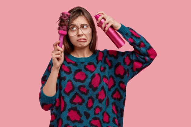 Indoor shot of displeased young female model has problems with hair, does hairstyle, combes with hairbrush, has frustrated look