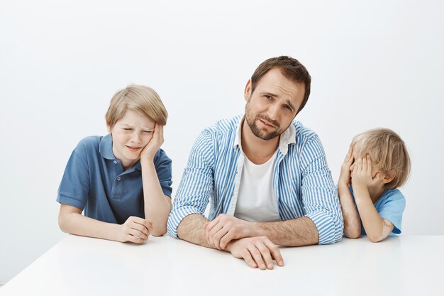 Indoor shot of displeased young sons and father sitting at table, whining and being upset, frowning from dislike, wanting eat meat while mom giving vegetables