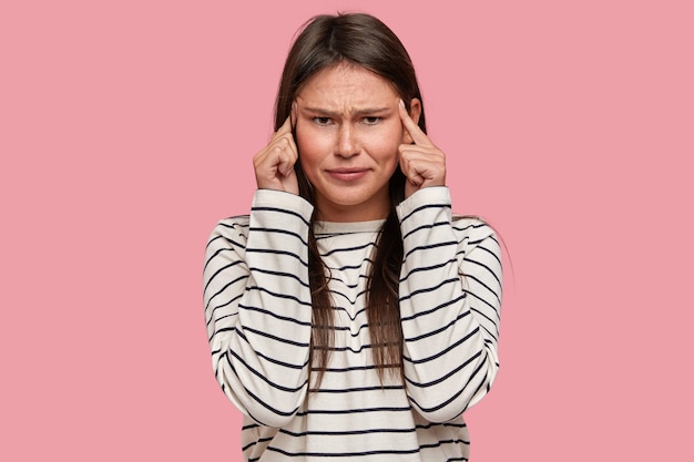 Indoor shot of disppleased young lady keeps both index fingers on temples, tries to recollect information