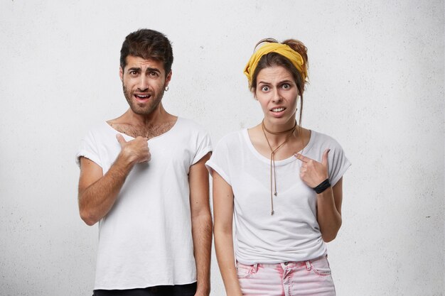 Indoor shot of dissatisfied male and female wearing casual clothes frowning their faces and pointing with fingers at themselves being puzzled to be chosen. European couple having confused look