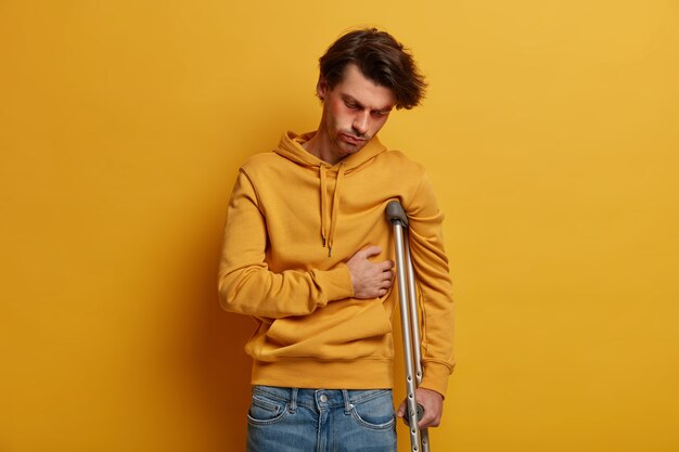 苦しんでいる男性の屋内ショットは、肋骨を骨折し、痛みに苦しみ、松葉杖の上に立って、道路で事故を起こし、黄色のスウェットシャツを着て、病気と怪我をして、黄色の壁を越えてポーズをとっています。モビリティエイド