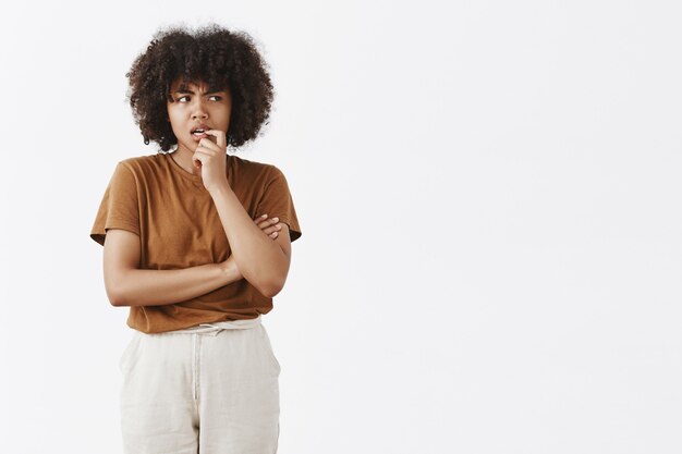 Снимок в помещении сомнительной и сомнительной молодой афро-американской женщины с афро-прической в коричневой футболке, кусающей ноготь и хмурящейся, смотрящей прямо во время принятия решения или размышлений