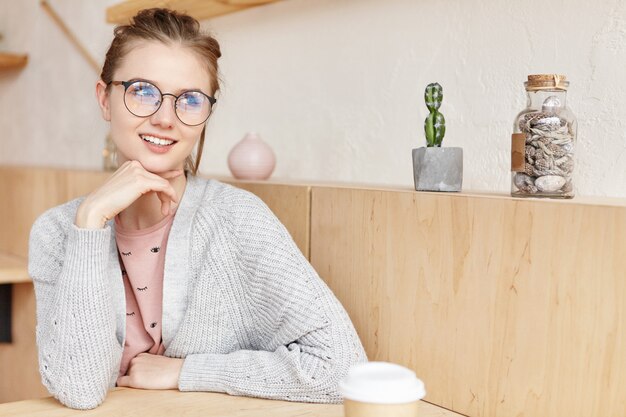 Indoor shot of dreamy adorable woman wearing round spectacles