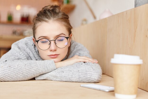 Indoor shot of dreamy adorable woman wearing round spectacles