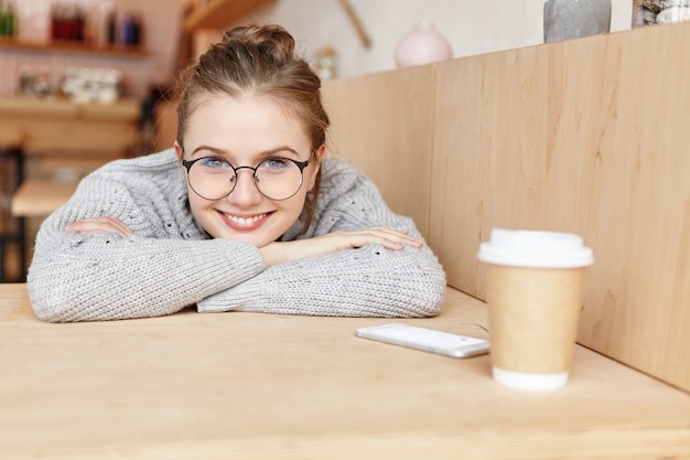 Indoor shot of dreamy adorable woman wearing round spectacles