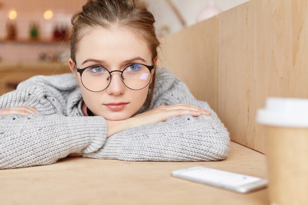 Indoor shot of dreamy adorable woman wearing round spectacles