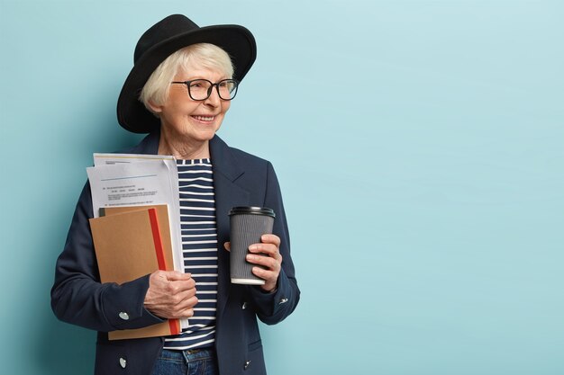 Indoor shot of elderly lady in high spirit, finishes paper work has coffee break, wears hat and jacket, looks aside, sees something pleasant. Experienced mature female teacher poses over blue wall