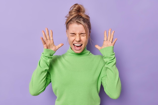 Free photo indoor shot of emotional annoyed young woman raises palms screams loudly dressed in casual green turtleneck feels irritated and bothered isolated over purple background human emotions concept