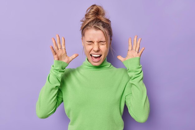 Indoor shot of emotional annoyed young woman raises palms screams loudly dressed in casual green turtleneck feels irritated and bothered isolated over purple background Human emotions concept