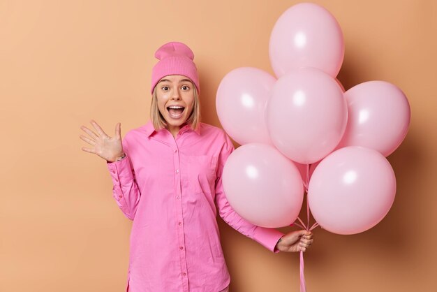 Indoor shot of emotional woman wears pink hat and shirt exclaims loudly holds bunch of helium balloons comes on birthday party isolated over beige background Festive event and celebration concept