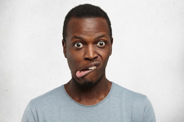 Внутренний снимок эмоционального молодого темнокожего мужчины в серой футболке, гримасничающего, выпучивая глаза