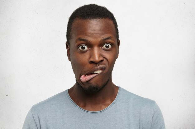 Indoor shot of emotional young dark-skinned man in grey t-shirt grimacing, bugging eyes out