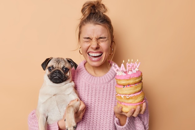 Indoor shot of emotional young woman exclaims loudly prepares for celebration of pets birthday holds pedigree pug dog and festive doughnuts with burning candles isolated over brown background