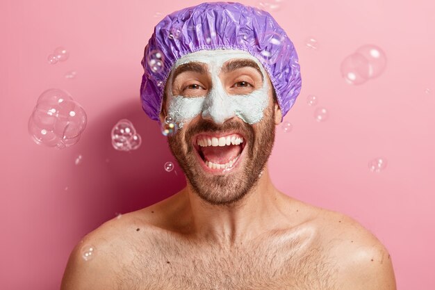 Indoor shot of emotive satisfied man with clay mask, enjoys taking shower and facial treatment, wears bathcap, soap bubbles flying around, washes body