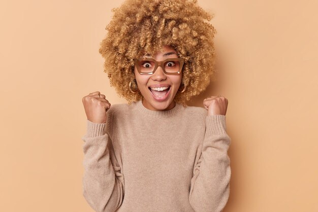 Indoor shot of excited emotional cheerful woman gestures with fists exclaims loudly from amazement being full of joy watches football game supports favorite game wears casual jumper isolated on beige