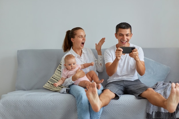 거실 소파에 앉아 있는 흥분한 가족의 실내 사진, 휴대전화를 손에 들고 있는 남편, 복권 당첨에 대한 훌륭한 소식, 유아를 안고 기뻐하는 사람들.