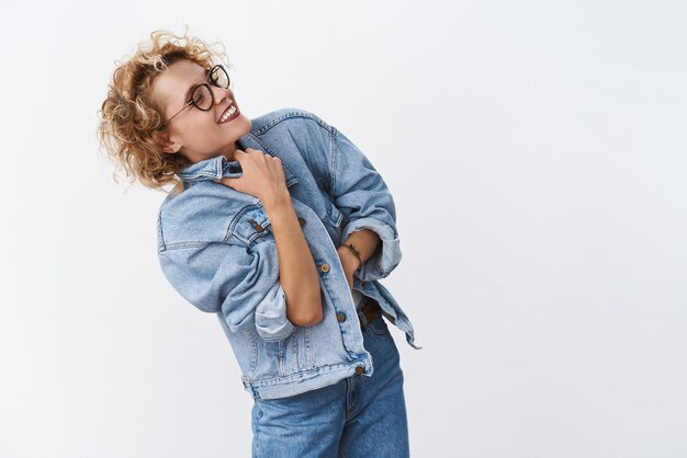 Indoor shot of excited happy and carefree stylish young european woman with shrot curly hairstyle turning right wearing glasses putting denim jacket on as feeling chilly having fun outdoors