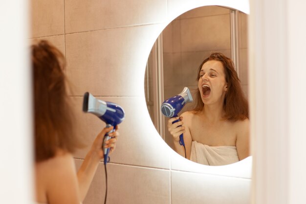 Indoor shot of excited happy young adult woman drying hair in bathroom with hair dryer, looking at reflection in the mirror and singing, keeps mouth opened.