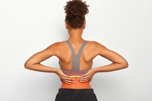 Indoor shot of faceless curly woman touches problematic zone of back, suffers from painful cramps, dressed in casual active wear, isolated over white background. Health care and medical concept
