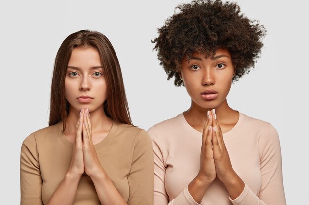Indoor shot of faithful mixed race women stand in praying gesture, ask for something desirable with serious expressions, keeps palms together
