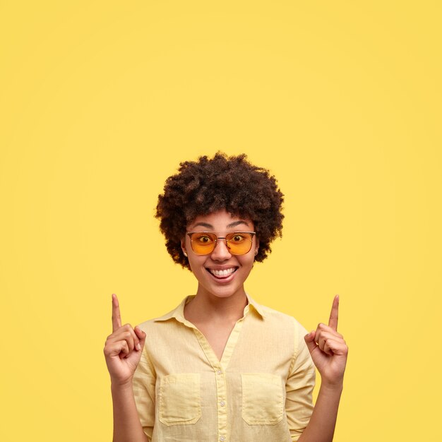 Indoor shot of fashionable black woman has afro hairstyle, wears trendy shades, shirt in one tone with wall, smiles positively, indicates upwards shows free space above head. use this for advert