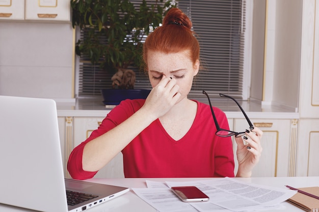 Indoor shot of fatigue overworked female entrepreneur takes off spectacles