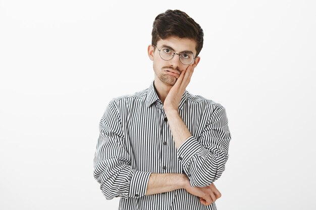 Indoor shot of fed up bored mature male model in striped shirt, holding palm on cheek and breathing out, staring with indifference and boredom, being annoyed with long office meeting