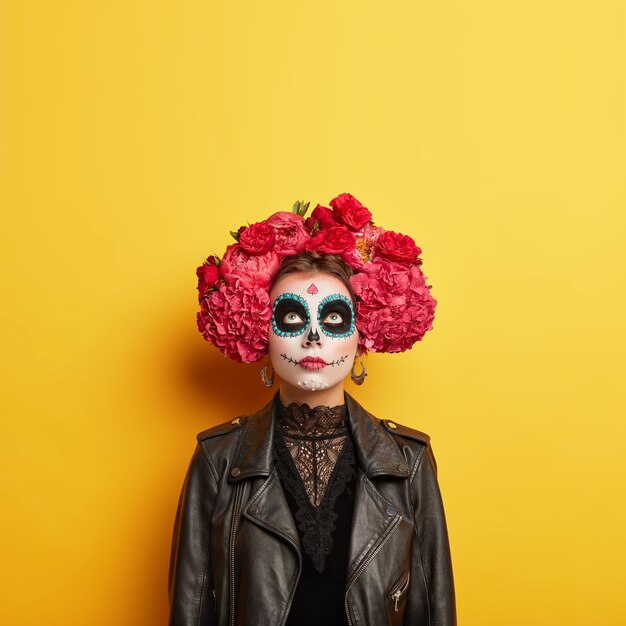 Indoor shot of female model has artistic designed face, wears professional horror makeup for Halloween holiday, dressed in special costume, focused upwards, isolated over yellow wall. Copy space