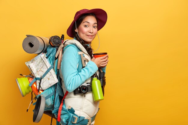 Indoor shot of female traveler has coffee break, enjoys trip, carries rucksack with necessary things and map, has long route, wears hat and comfortable clothing