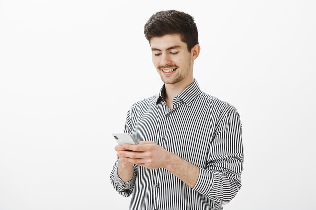 Indoor shot of focused satisfied attractive man with beard and moustache, holding smartphone, looking at screen with pleased happy smile, messaging lawer about divorce