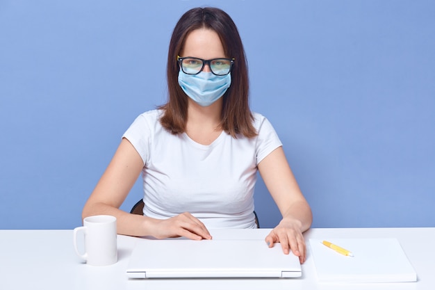 Free photo indoor shot of freelancer accountant working at hone, lady wearing casual white t shirt, eyeglasses and medical mask