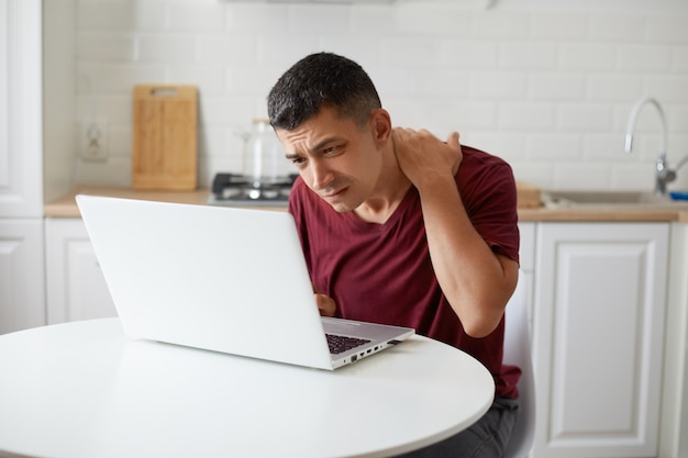 Indoor shot of freelancer male worker sitting in kithen at table in front of white notebook, looking intently at laptop display trying to notice importaint thing, keeps hand on neck.