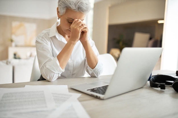 Indoor shot of frustrated unhappy middle aged businesswoman managing papers while sitting at office desk in front of open laptop, looking down with hands on her face