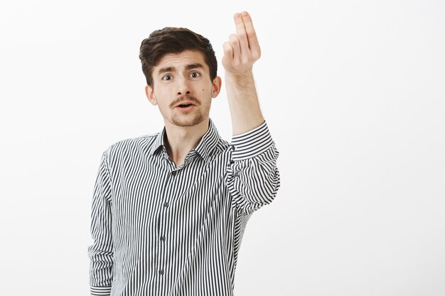 Indoor shot of funny ordinary european male with moustache and beard, talking passionately while raising hand with italian gesture