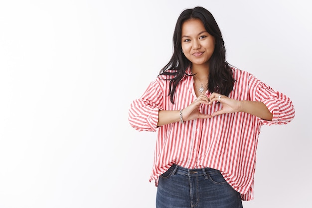 Indoor shot of girlfriend in striped blouse with tattoo confessing in sympathy showing heart gesture near body and smiling cute at camera, sharing love and romantic feelings. Relationship concept