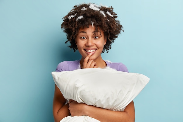 Indoor shot of glad dark skinned woman holds soft white pillow, sleeps well at night, enjoys rest, comfort and leisure, has cheerful expression, stands over blue wall. People, awakening concept