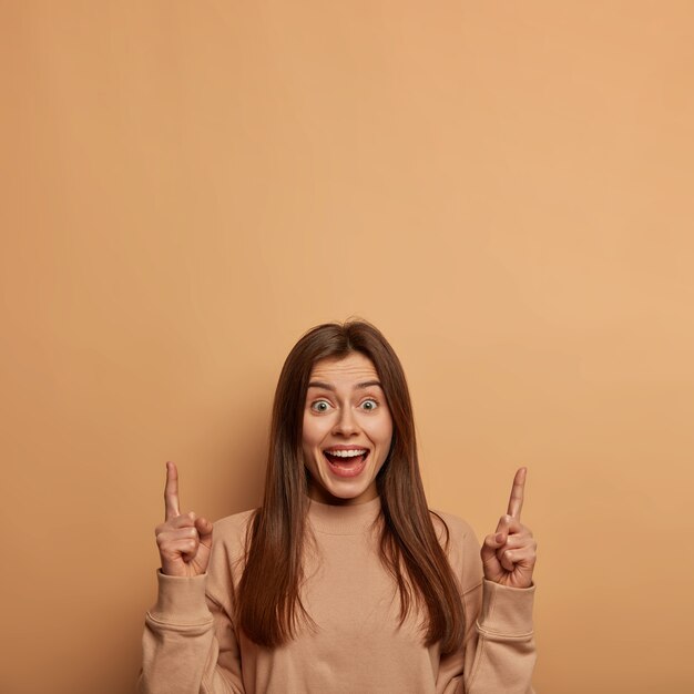 Indoor shot of glad delighted woman points above with both index fingers, laughs joyfully, advertises cool blank space, wears loose sweater