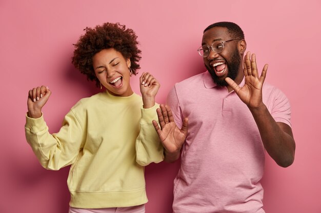 Indoor shot of glad girlfriend and boyfriend dance hip hop with joy, celebrate something, laugh and move with hands up, wear spectacles, jumper and t shirt