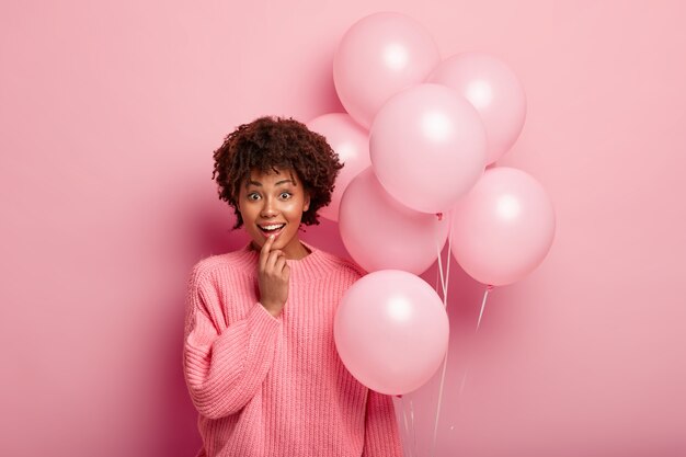 Indoor shot of glad optimistic young Afro American surprised to get present, holds air balloons, wears oversized jumper, keeps finger on lower lip, satisfied to be present on party celebration.