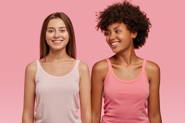 Free photo indoor shot of glad satisfied mixed race sisters being in high spirit, look happily, rejoice weekend and pastime spending together, isolated over pink wall. positive feelings concept.