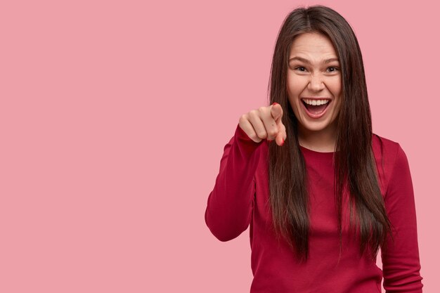 Indoor shot of glad young European woman smiles gladfully, points at camera with index finger, chooses you