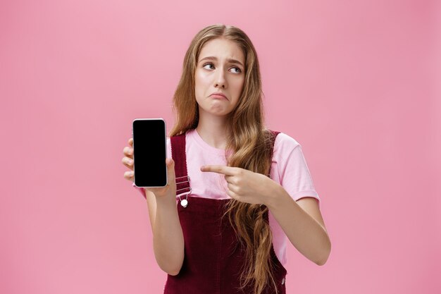 우울하고 실망한 귀여운 어린 소녀가 셀을 가리키는 스마트폰을 들고 있는 실내 사진