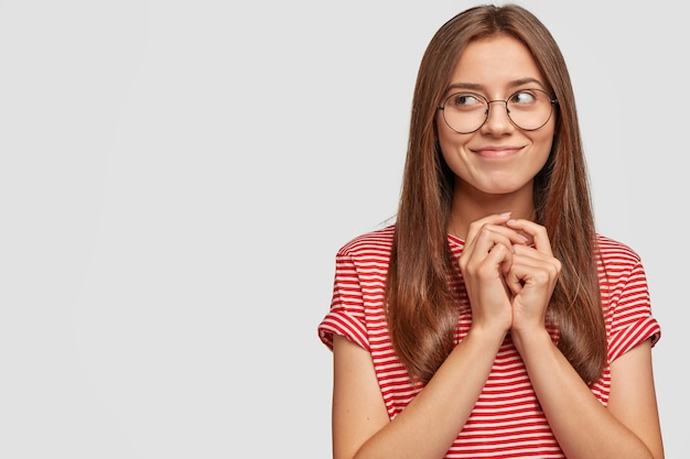 Indoor shot of good looking brunette woman with satisfied expression, looks in intrigue aside, keeps hands together