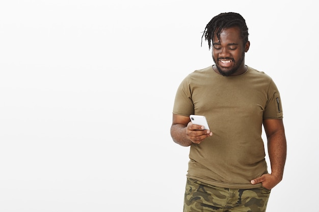 Красивый счастливый парень в коричневой футболке позирует на фоне белой стены со своим телефоном в помещении