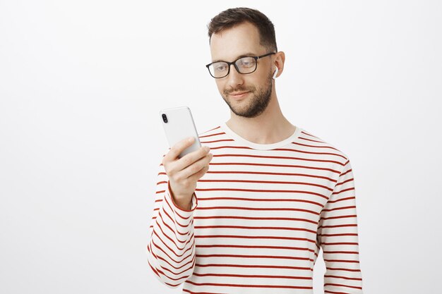 Indoor shot of good-looking relaxed european man in glasses, holding smartphone, picking song to listen, wearing wireless earphone