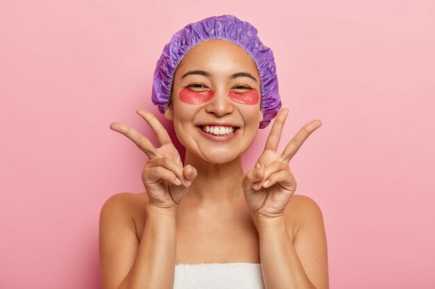 Indoor shot of good looking smiling Asian girl make peace gesture with both hands, enjoys uder eye treatment, applies collagen patches, visits cosmetologist wears shower cap on head. Face care concept