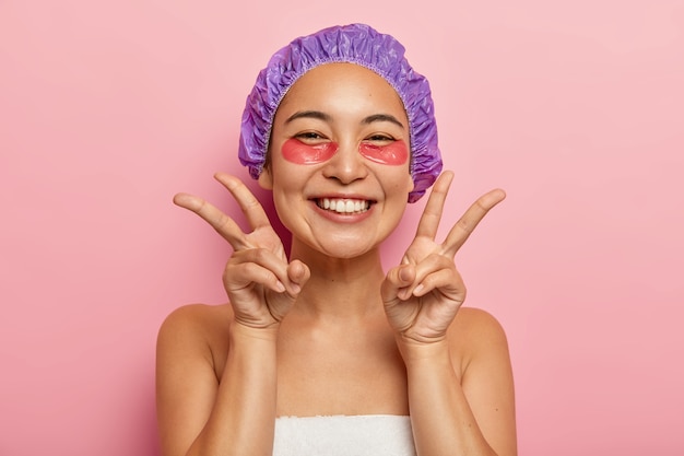 格好良い笑顔のアジアの女の子の屋内ショットは、両手で平和のジェスチャーをし、目の治療を楽しんで、コラーゲンパッチを適用し、美容師を訪問し、頭にシャワーキャップを着用します。フェイスケアのコンセプト