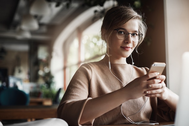 Indoor shot of good-looking stylish and happy caucasian female with fair hair in glasses, holding smartphone and wearing earbuds while watching video, distracting to look and smile