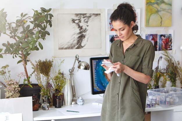 Indoor shot of gorgeous beautiful brunette young woman designer typing text message on mobile phone, shopping online, ordering paint, canvas or frame. People, art, creativity and technology concept