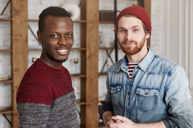 Indoor shot of handsome African male in cozy sweater standing next to his bearded hipster friend